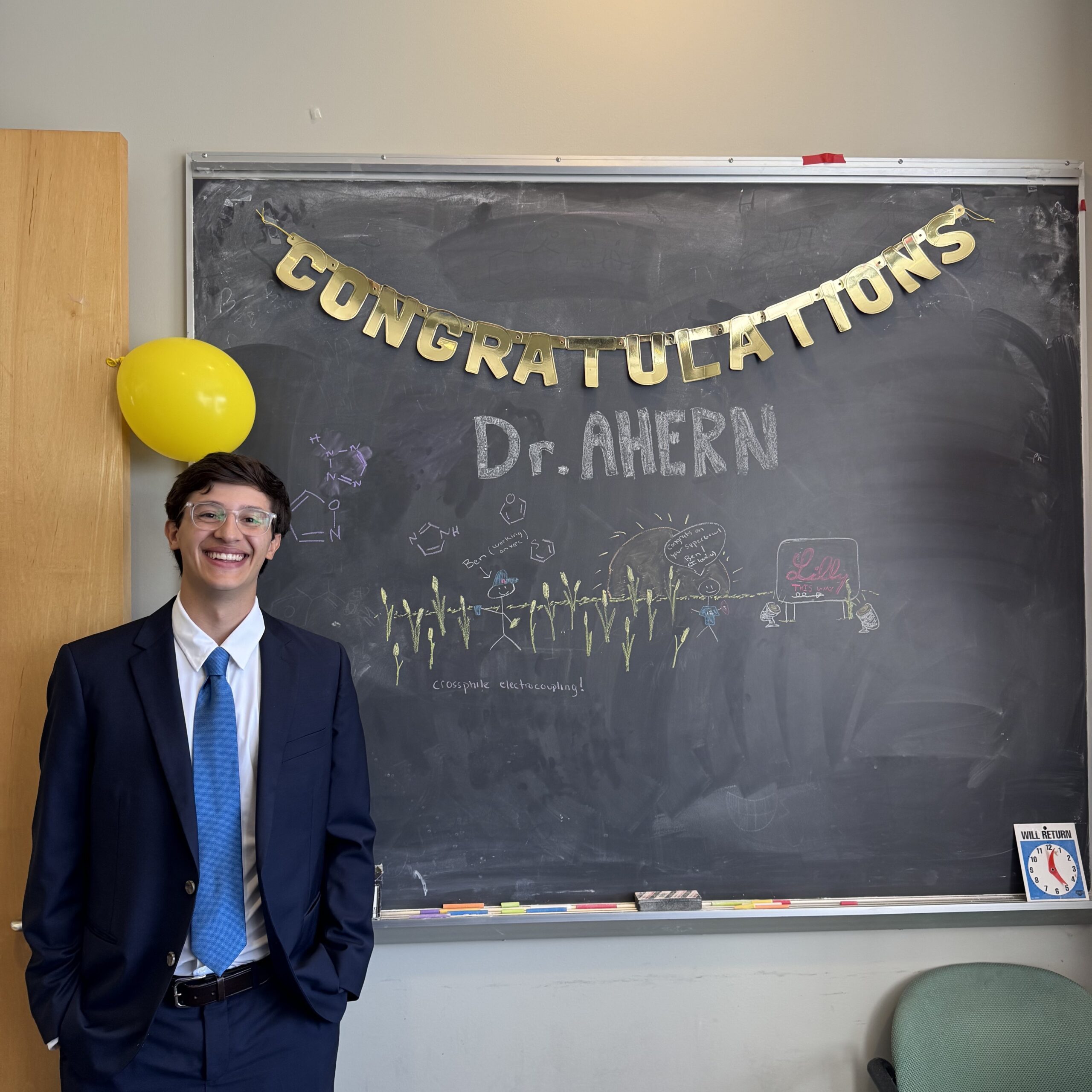Ben Ahern standing in front of a chalkboard that says "Congratulations Dr. Ahern"